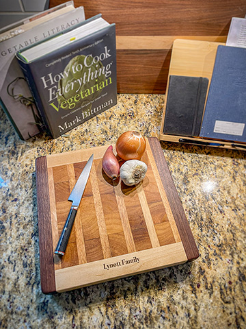 Striped maple, walnut and cherry edge-grain cutting board branded for the Lynott family, styled with onions, garlic, a chef's knife, and a cookbook