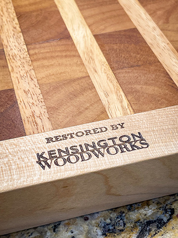 Macro detail of a restored striped cutting board branded with a burned-in mark reading RESTORED BY KENSINGTON WOODWORKS