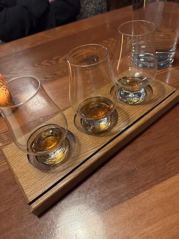 A wooden whisky tasting flight tray with three Glencairn glasses set into recessed circles, photographed on a bar in low light